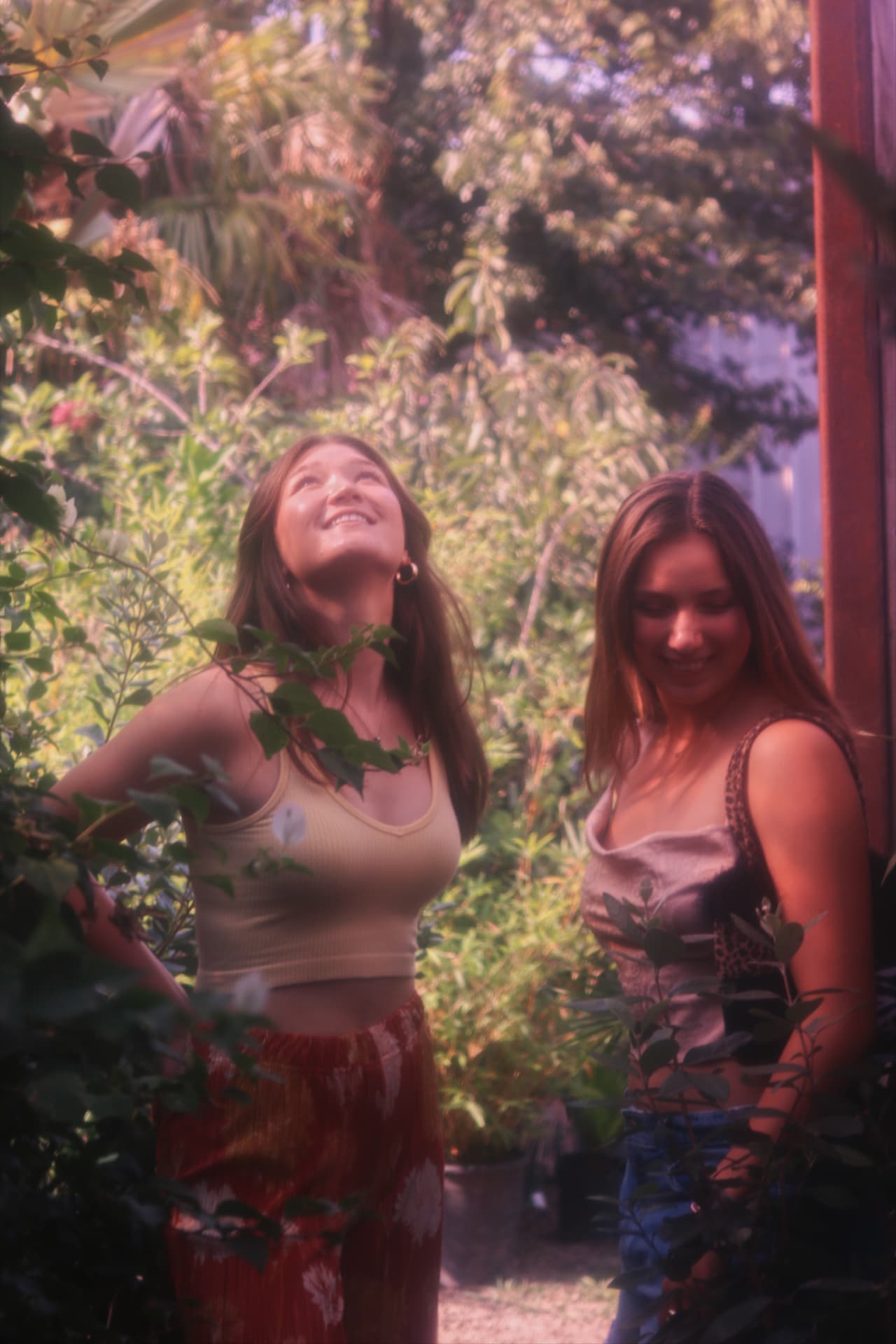 Two women laughing among lush garden greenery in warm light