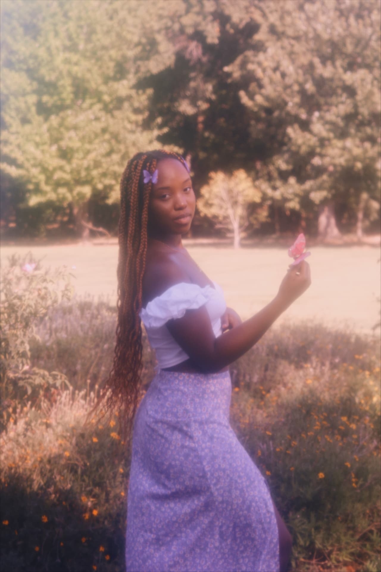A woman with butterfly clips in her braids holding a pink butterfly in a sunlit meadow