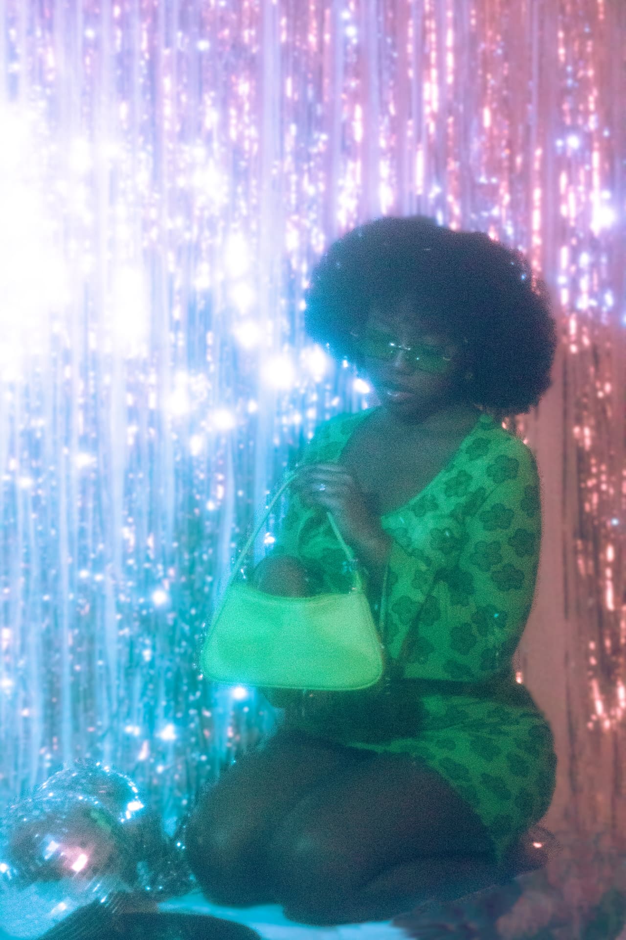 A crouching woman in neon green before a tinsel backdrop, a disco ball low at her side