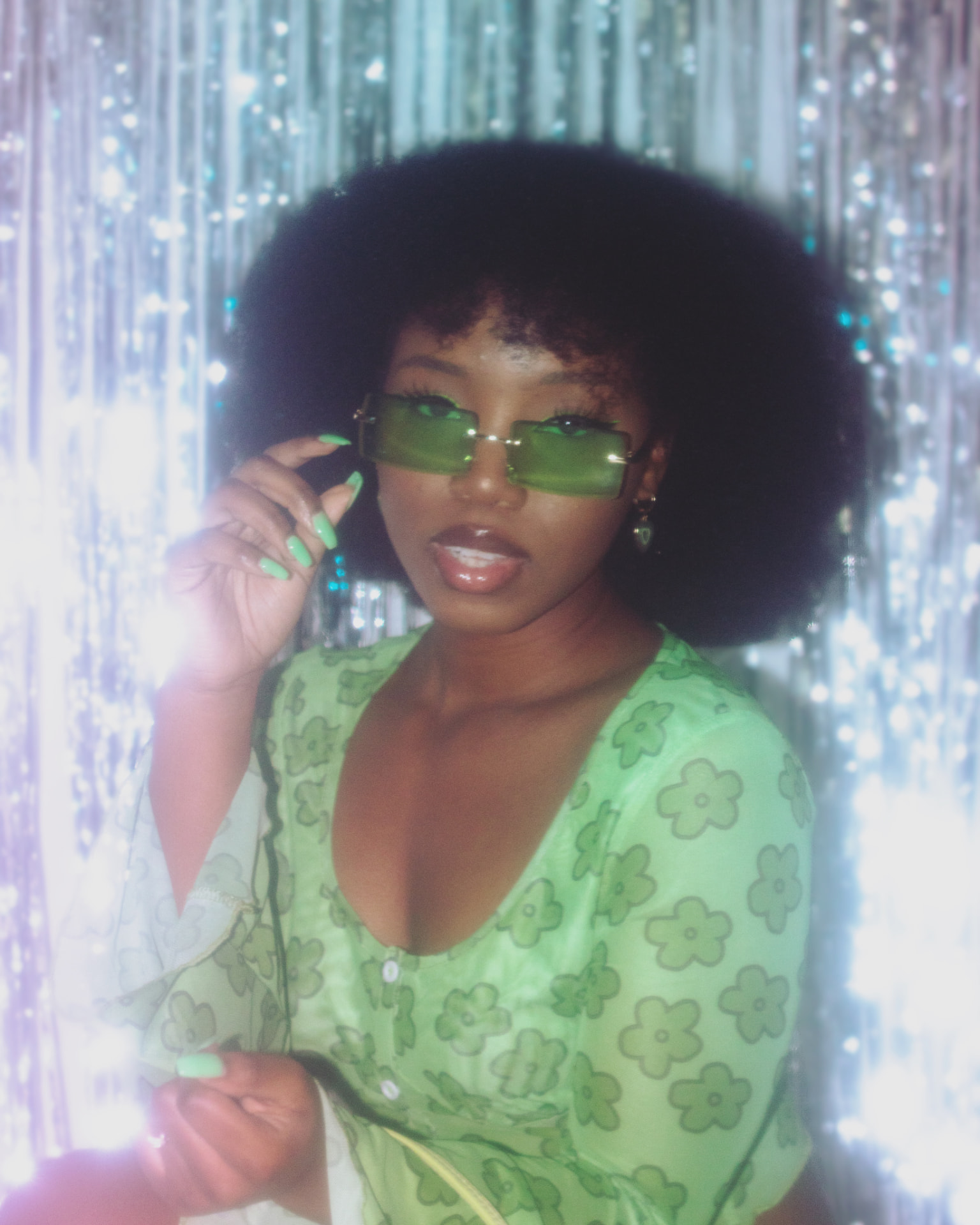 Close portrait of a woman in lime-green sunglasses before a shimmering tinsel curtain