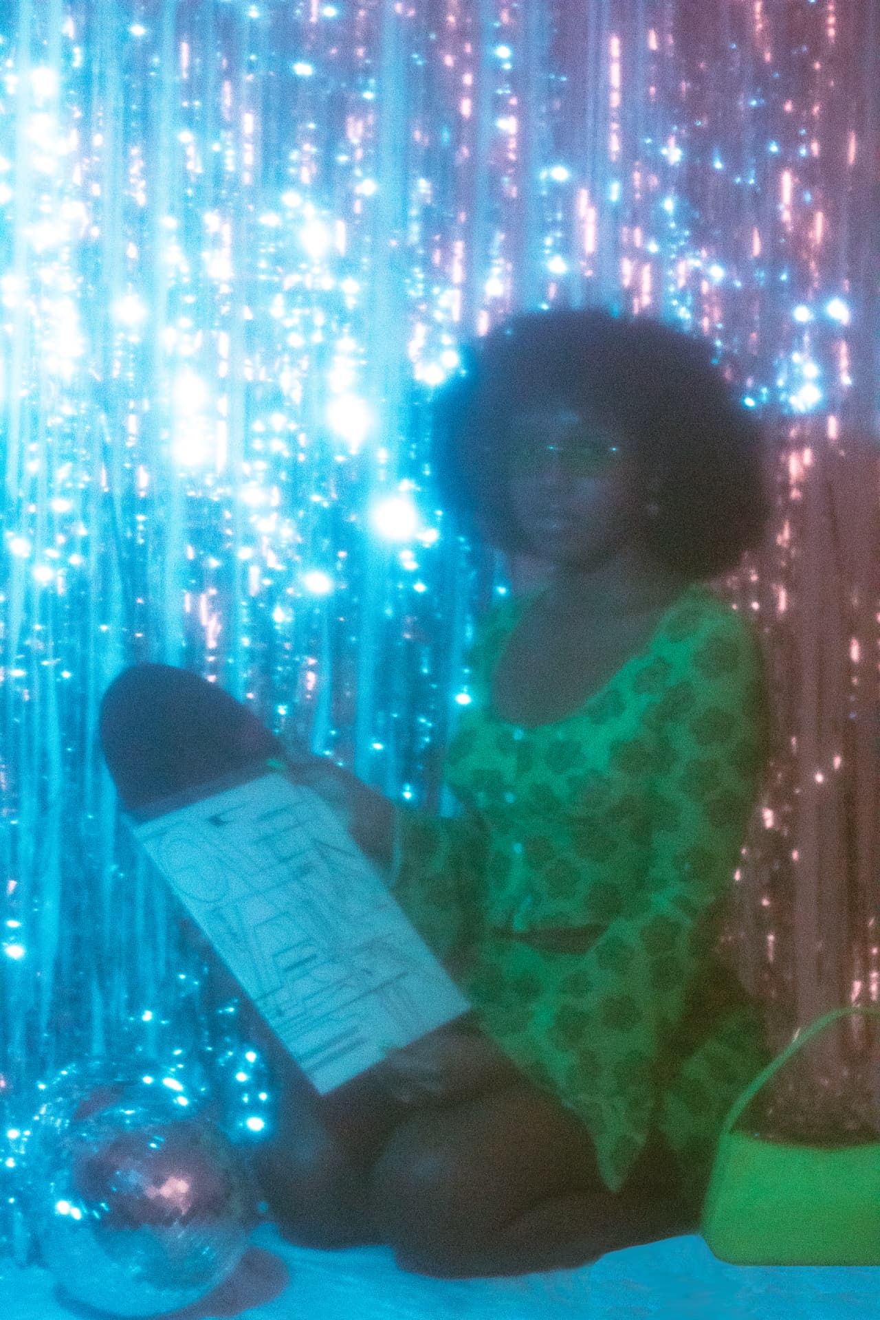 A crouching woman holding a record sleeve, bathed in cyan flare before a tinsel curtain
